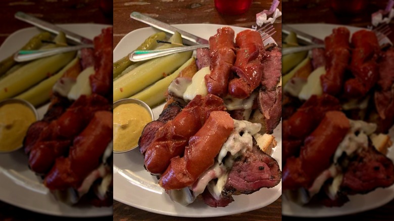 Open-faced pastrami topped with knockwurst on a platter with pickles and mustard