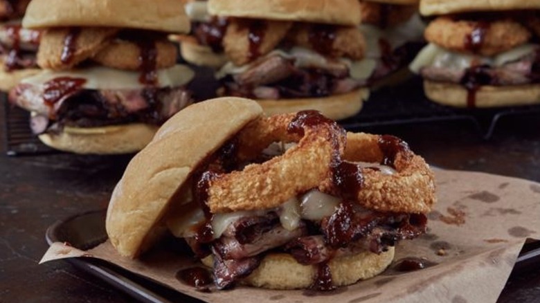 Sauce, cheese, and onion rings topping brisket on a roll served in front of like sandwiches