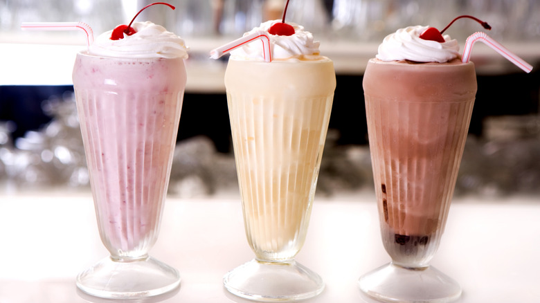 Three milkshakes in vanilla, strawberry, and chocolate are in classic diner glasses, topped with whipped cream and cherries, with pin-stripe straws