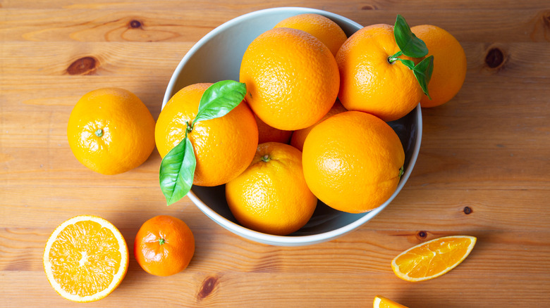 Several oranges in a bowl, with others, including cut pieces, around it