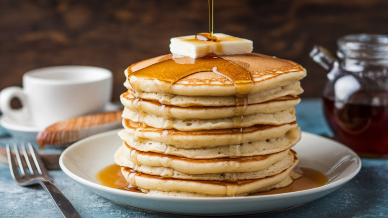 Stack of pancakes with cube of butter and drizzling maple syrup white plate with a white coffee cup and jar of maple syrup in background