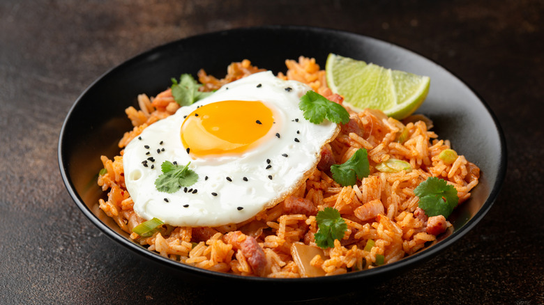 Kimchi fried rice with bacon, eggs, lime, and garnish in a black bowl, on a dark table.