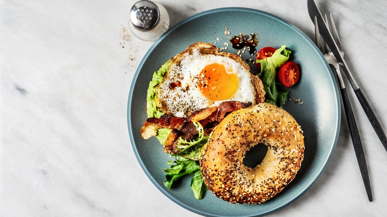 Top view of a breakfast sandwich on an everything bagel, with egg, bacon, avocado, lettuce, and various seasonings on a marble table with utensils.