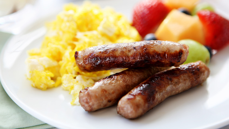 Three cooked breakfast sausage links on a white plate with scrambled eggs and fresh fruit salad