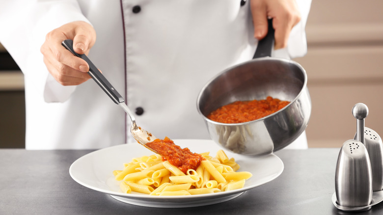 Person wearing a white chef's coat spooning sauce from a pot onto pasta on a plate