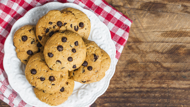 A plate of soft-looking chocolate chip cookies sit on a white plate on a checkered towel