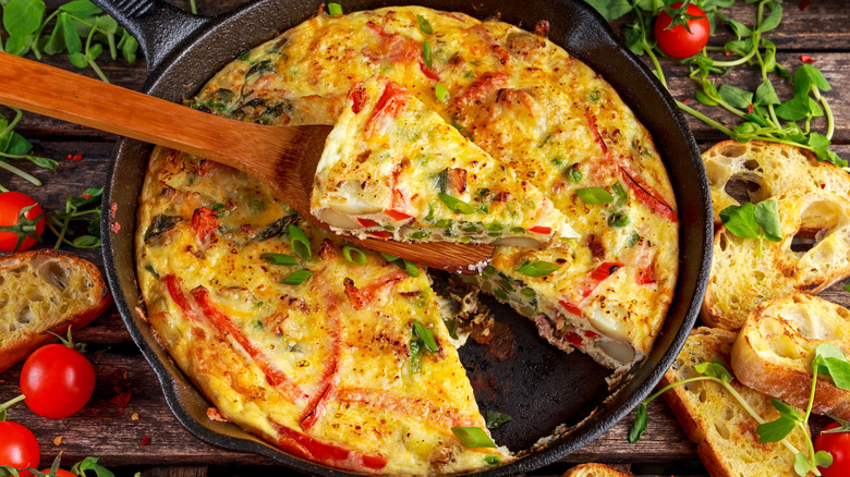 Vegetable frittata in a black cast iron pan with one slice cut out on a wooden spoon, surrounded by slices of Italian bread, cherry tomatoes, and herbs