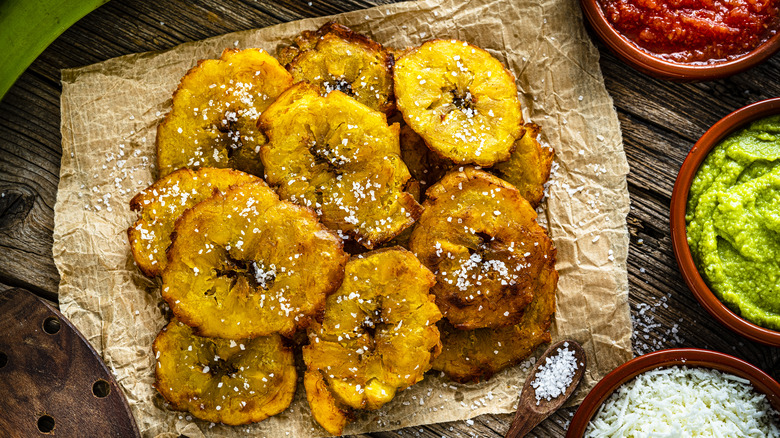Tostones on a wooden board next to seasonings and sauces.