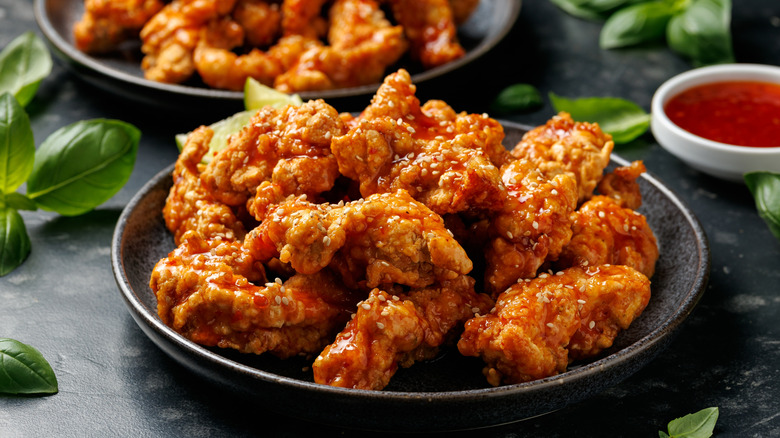 A plate of Korean fried chicken is topped with sesame seeds and surrounded by fresh basil leaves.