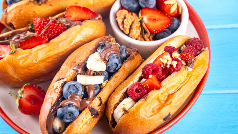 Two plates of hot dog buns filled with berries and fruits and chocolate sauce on a painted blue wooden table