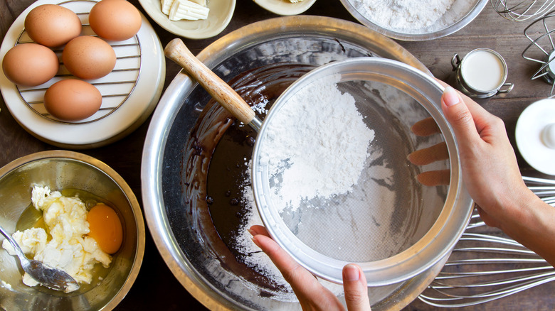 Person sifting flour in a bowl with eggs on the side
