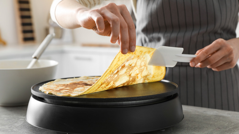 A woman wearing an apron flips a crepe on a single burner with a rubber spatula.
