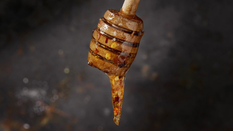 A honey dipper held aloft drips honey flecked with red pepper flakes against a dark background