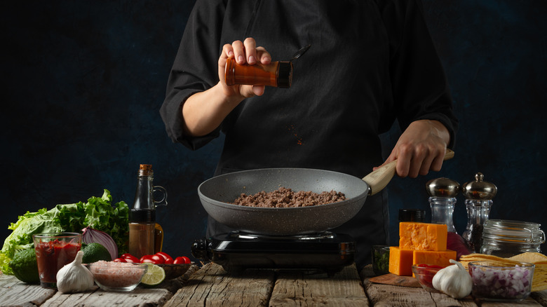 A chef cooking ground beef in a skillet sprinkles in seasoning