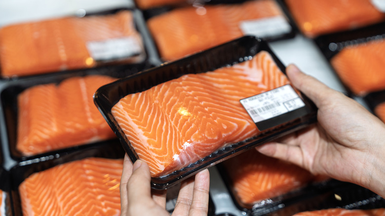 A person is holding a tray of packaged salmon over a seafood display case in a grocery store.