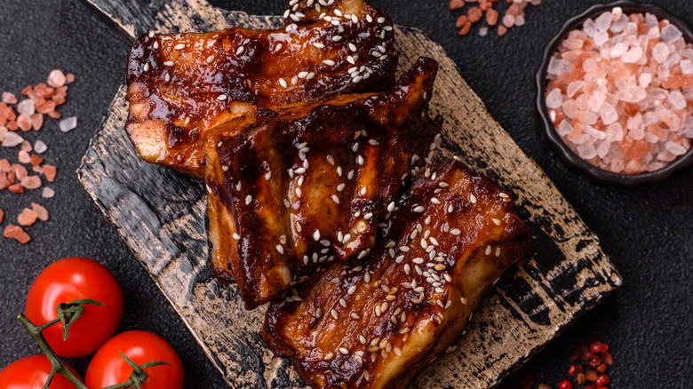 Glazed ribs like on a wooden serving board beside tomatoes and coarse pink salt