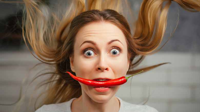 A person with long blonde hair blowing holds a chili pepper in their mouth