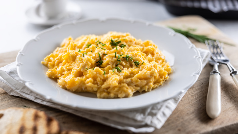 Fluffy scrambled eggs with sprinkled chives on a round white plate with a knife and fork next to it