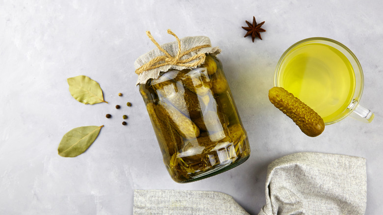 Jar of pickles on its side next to a clear glass cup with pickle juice and a small gherkin on it, and a few scattered whole spices on a gray background
