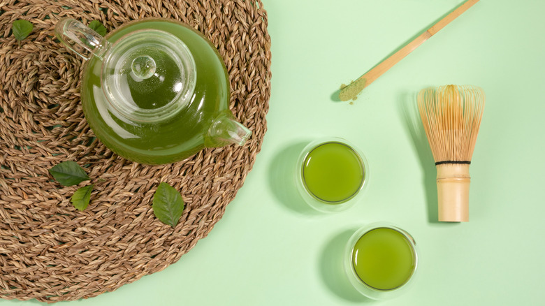 Two cups of matcha with a whisk and spoon next to a matcha cup on a woven placemat