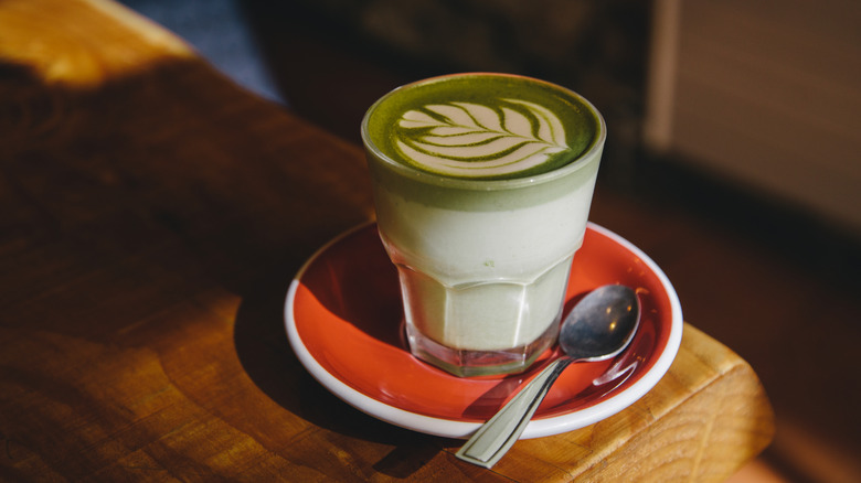 Matcha latte with latte art on a red saucer with spoon on a wooden table