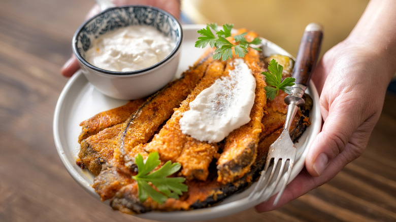 Hands holding a plate of eggplant fries topped with creamy spread, and a side bowl of dipping yogurt sauce. The person is wearing a yellow shirt and standing on a wooden floor.
