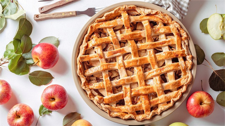 Fully-baked apple pie with lattice crust on a white background surrounded by whole apples