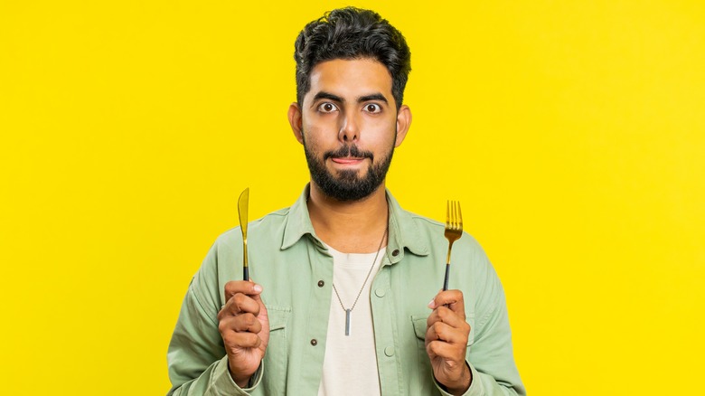 Man in green shirt before a yellow background licks lips while holding knife and fork