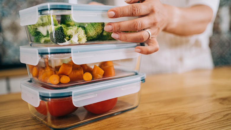 A female hand secures the lid on a reusable glass food container holding broccoli, which is stacked on two other glass containers containing carrots and tomatoes