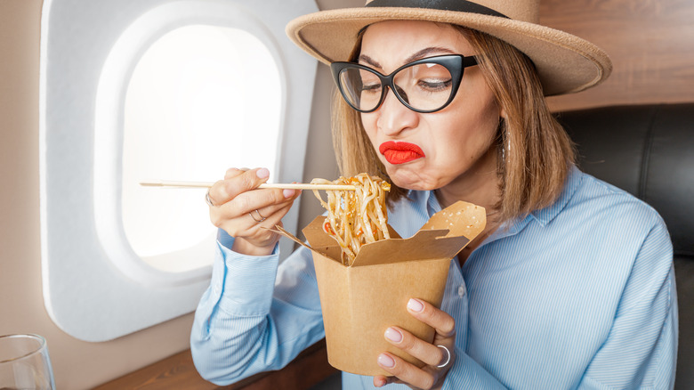 A woman eating a box of noodles on a plane