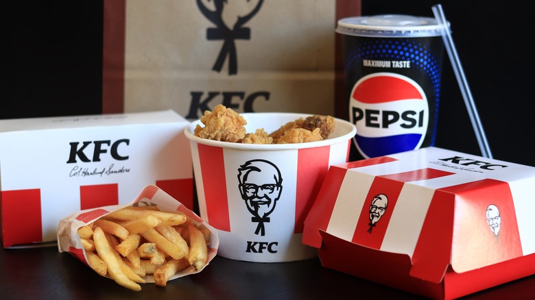 KFC food, including a bucket of fried chicken, french fries, a soda, two closed food container boxes, and a KFC paper bag