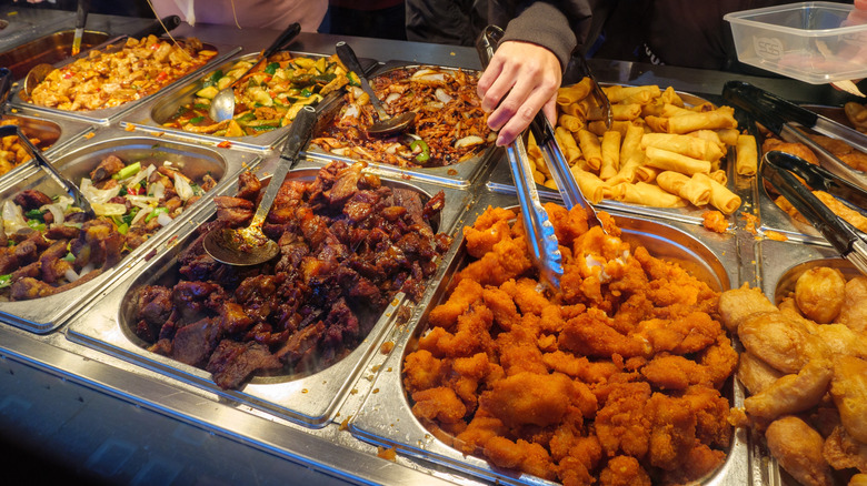 Hands holding tongs retrieve fried chicken pieces from a buffet serving dish