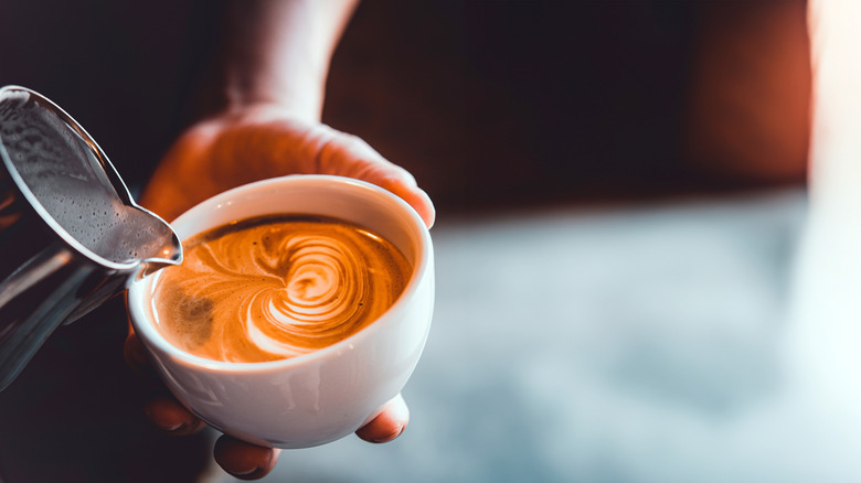 Close up of barista pouring a café au lait from a frothing pitcher into a porcelain bowl. Blurred, nondescript background.