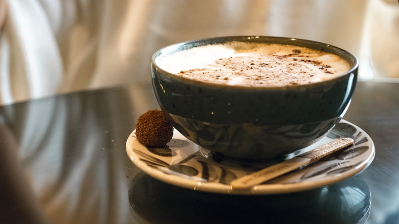 French café au lait in a green, porcelain bowl on a saucer. A single chocolate truffle and stir stick are sitting next to the bowl, all on a dark table and blurred background.