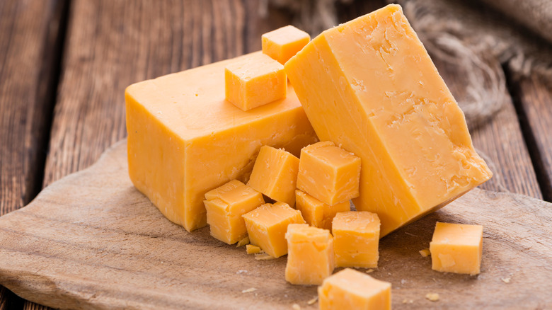 A block of orange cheddar cheese on a wooden surface next to smaller cubes of cheddar