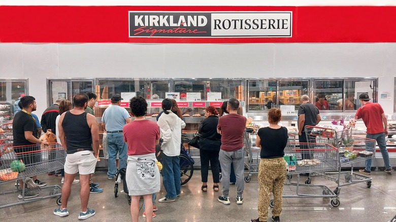 A large ground of costco shoppers waits by a Rotisserie chicken aisle
