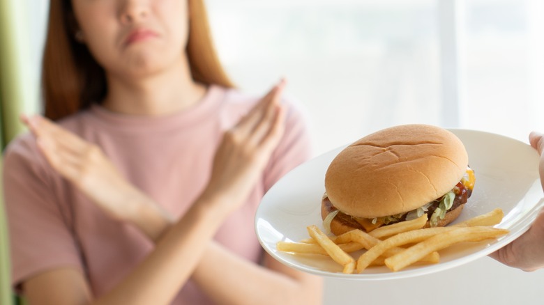 A frowning woman forms an X with her arms in refusal of a plate with a fast-food hamburger and fries being offered to her
