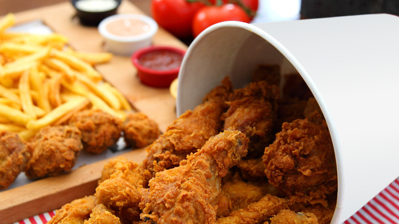 A bucket of fried chicken spilled over a red and white striped tablecloth with fries and chicken nuggets in the background