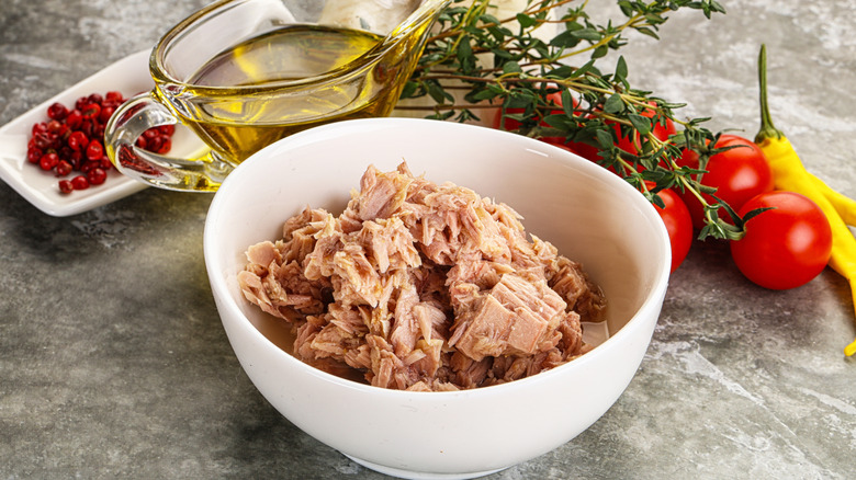 Canned tuna in a white bowl sits on a counter surrounded by tomatoes, peppers, herbs, a glass container of oil, and pink peppercorns on a white dish