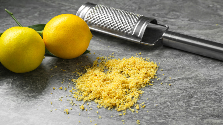 Two lemons sit on a counter near a metal lemon zester and a pile of zest