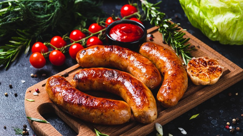 Smoked sausages sit on a wooden cutting board surrounded by ingredients like garlic, tomatoes, and rosemary sprigs