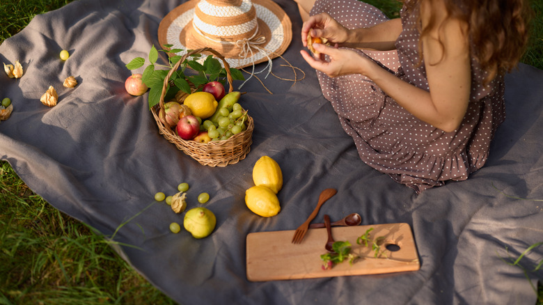 A woman sits on a blanket with a fruit-filled basket, hat, serving board, and utensils