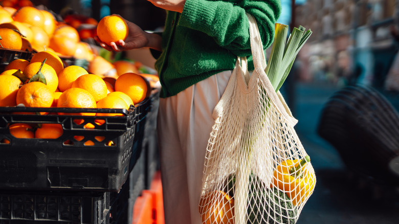 Woman with mesh bag of produce chooses oranges from a stand