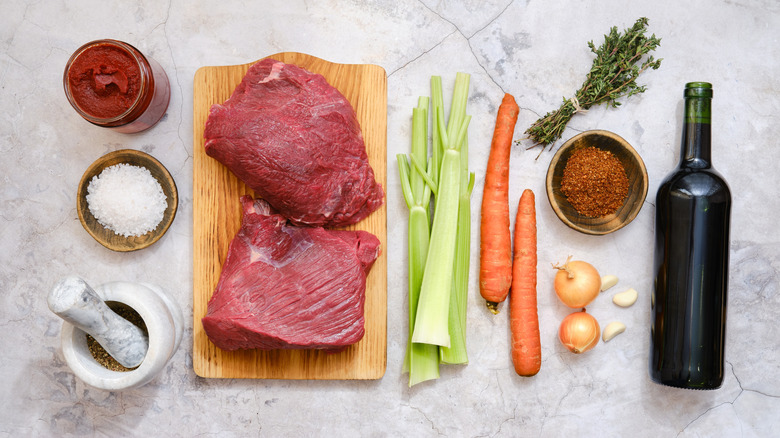 Beef stew ingredients including meat, carrots, celery, onions, red wine, and spices on a white surface