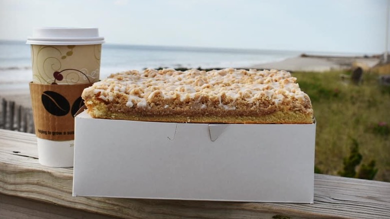 Crumb cake in box on boardwalk railing beside a cup of coffee