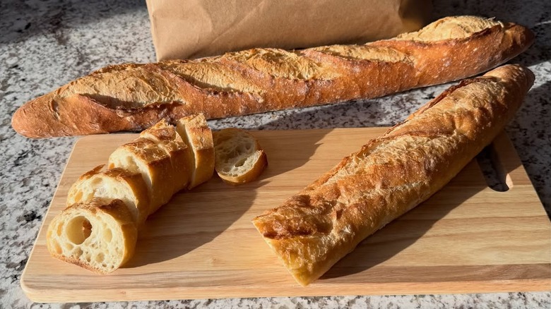 one full french baguette and one baguette cut in half, one cut into slices from On the Rise Artisan Breads, laid out on a cutting board atop a marble counter