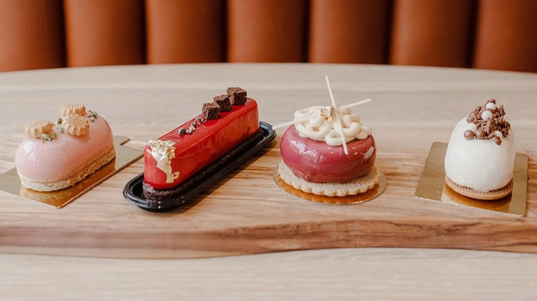 A wooden tray sitting on the table of a booth, displaying four complex, artful pastries in colors of red, pink, brown, and white