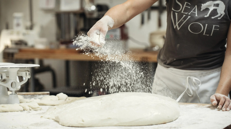 A baker at Sea Wolf Bakery (wearing a Sea Wolf Bakery t-shirt and a white apron) preparing bread dough with flour in the bakery's kitchen