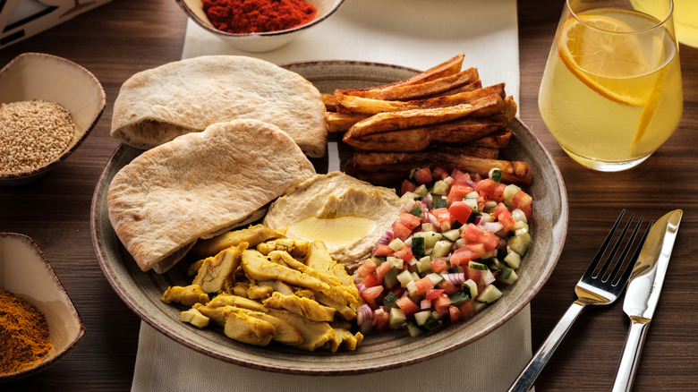 A plate has rustic fries, Israeli salad, chicken shawarma, hummus, and pita bread, and sits next to a glass of lemon drink as well as bowls of spices