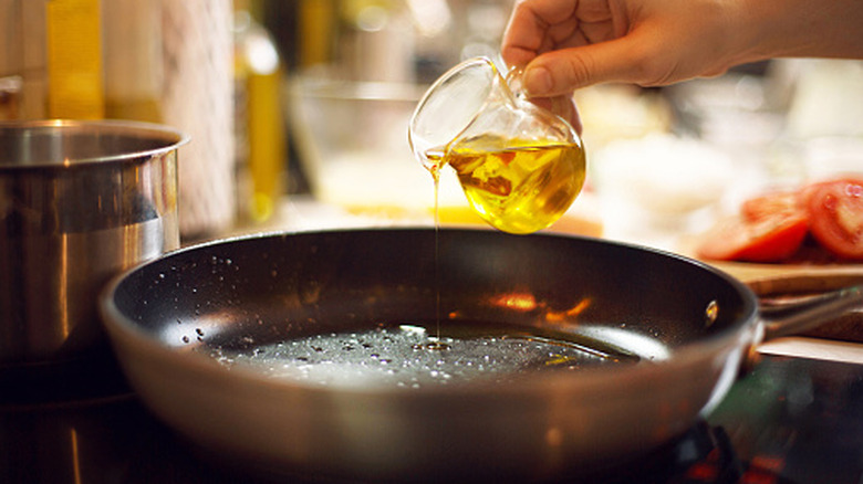 A hand pouring oil from a small glass pitcher onto a skillet.